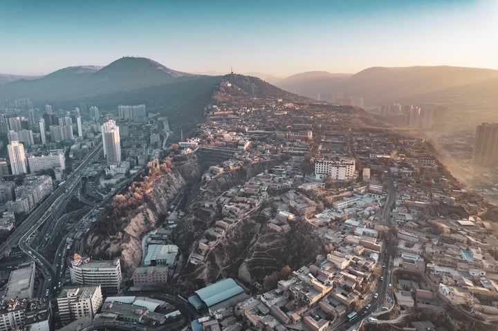 An aerial view of a mountainous city with a hillside, densely populated with buildings of varying heights, surrounded by greenery. The city is situated on a hill, with roads and highways running through it. The sun casts a warm glow on the city, highlighting its architectural details.