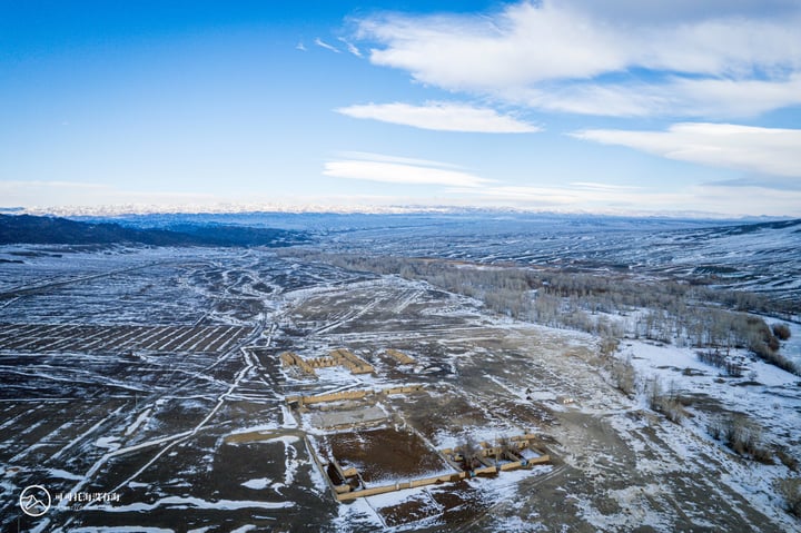 An aerial view of a snowy landscape with a large open field, a few scattered buildings, and a mountain range in the distance.