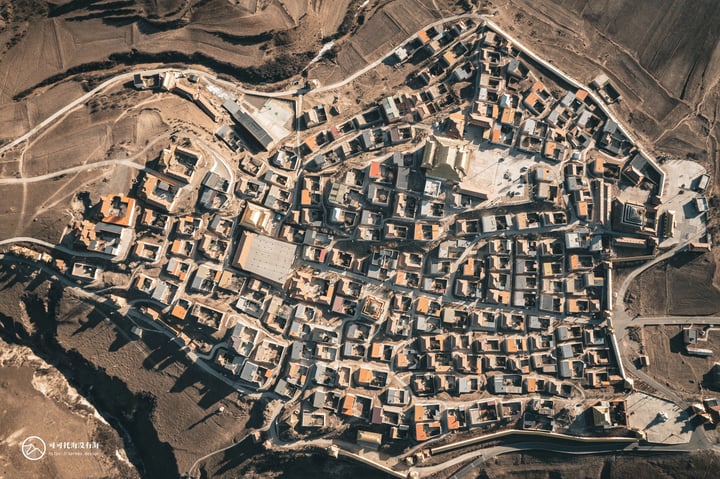 An aerial view of a village with a mix of orange and brown buildings, surrounded by a network of roads and fields, with a mountain range in the background.