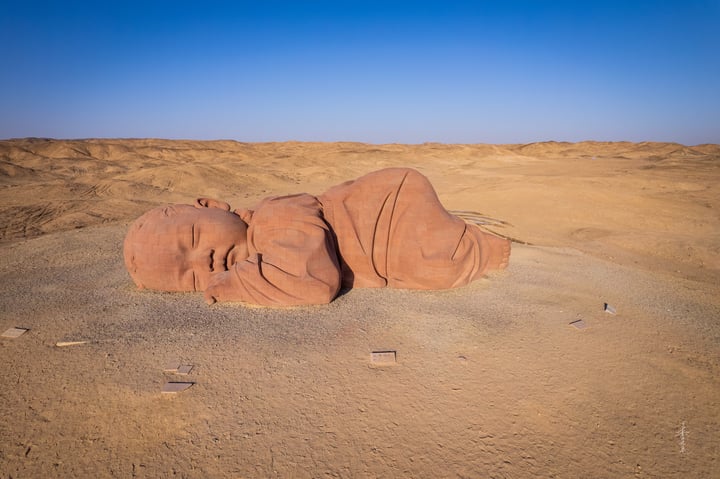 A large, brown, clay sculpture of a baby lying on the ground in a desert-like environment, with a few people in the distance.