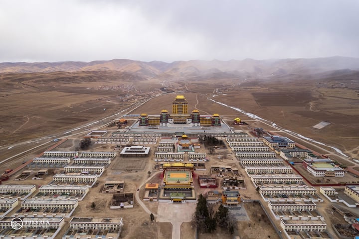 An aerial view of a large, white and yellow Buddhist temple with a golden dome, surrounded by a barren landscape of mountains and hills, under a cloudy sky.