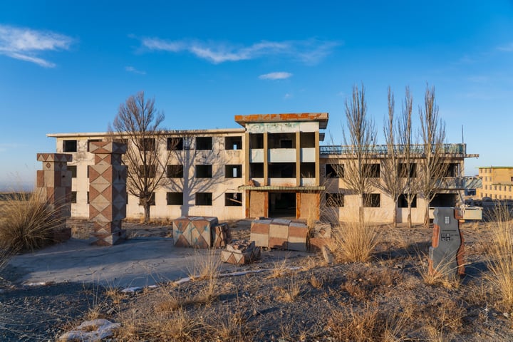 An abandoned, two-story building with a concrete base and a brick facade stands in a barren landscape, surrounded by dry grass and shrubs. The sky is clear blue, and the building's rusted metal structure contrasts with the surrounding environment.