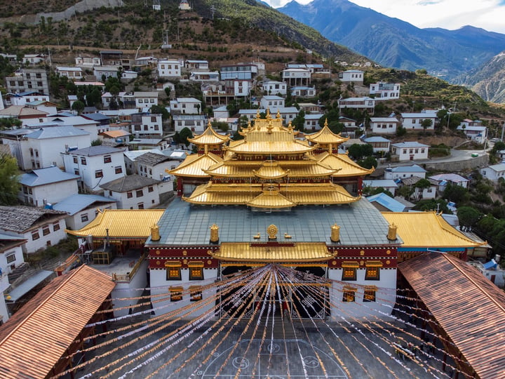 The image features the Lhasay Monastery in Nepal, a golden temple with a red roof and intricate designs, surrounded by white buildings and mountains. The perspective is from above, showcasing the temple's grandeur and the surrounding landscape.