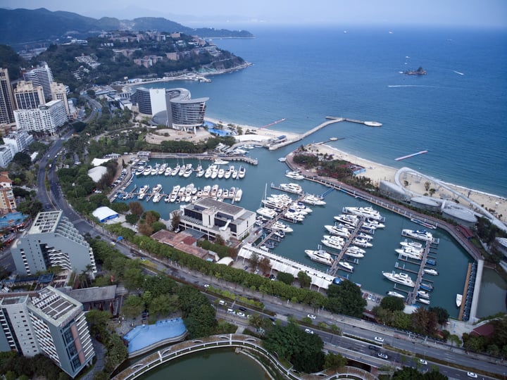 An aerial view of a bustling harbor with numerous boats docked in calm waters, surrounded by a cityscape of tall buildings and a mountain range in the background.