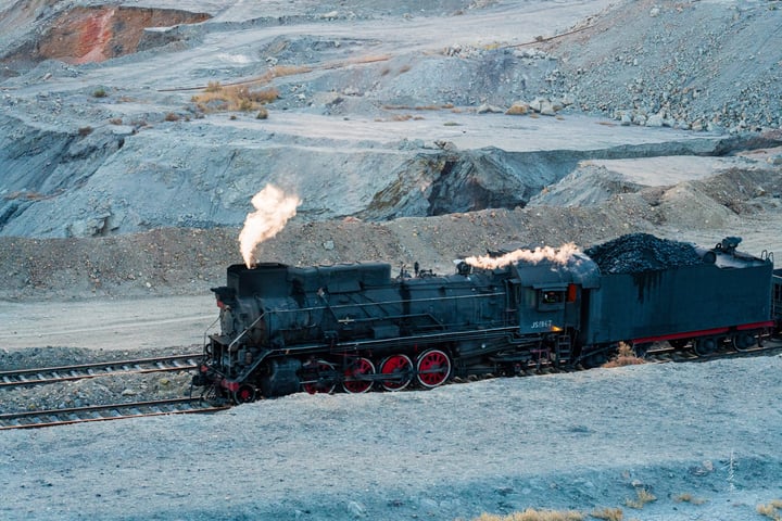 A black steam locomotive with red wheels is traveling on a track in a desert-like area, emitting white smoke.