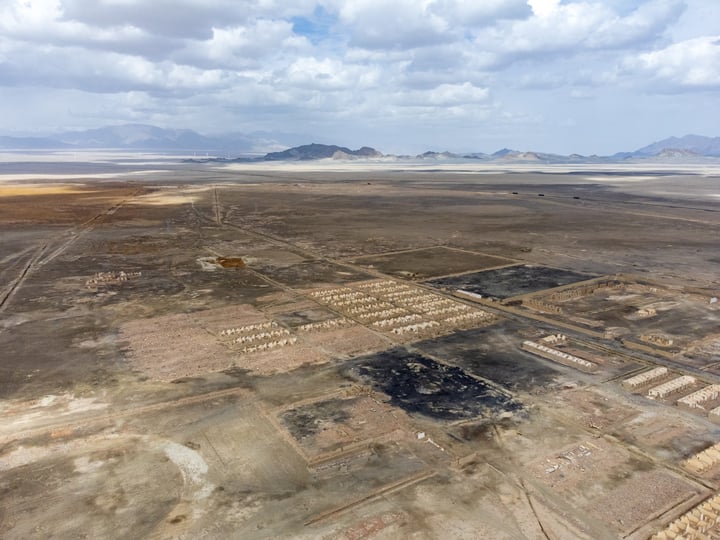 An aerial view of a vast, barren desert landscape with scattered dirt patches and mountains in the distance.