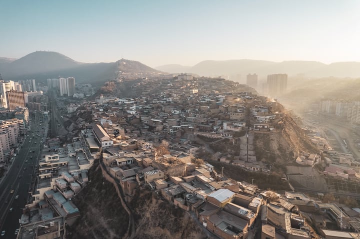 An aerial view of a mountainous city with a hillside, densely populated with buildings of varying heights, surrounded by greenery. The city is situated on a hill, with roads and highways running through it. The sun casts a warm glow on the city, highlighting its architectural details.