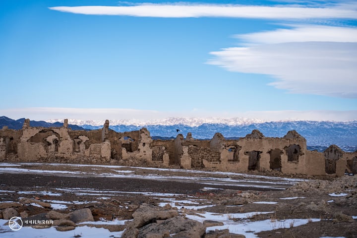 A desolate, weathered building with a single window stands in a barren landscape, surrounded by snow-covered mountains under a clear blue sky.