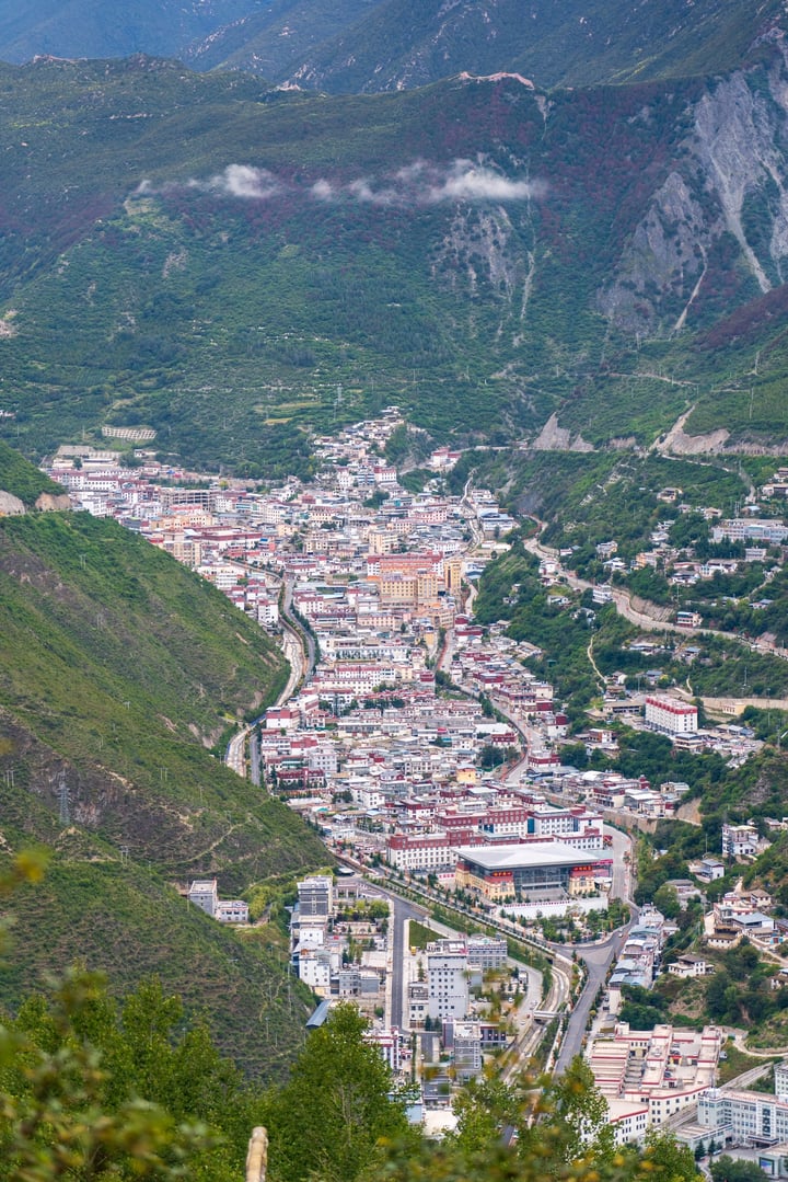 An aerial view of a Deqin County, surrounded by lush greenery and surrounded by mountains. The town is densely populated with houses and buildings, and the roads crisscross the valley. The town is surrounded by trees and hills, creating a serene and picturesque scene.