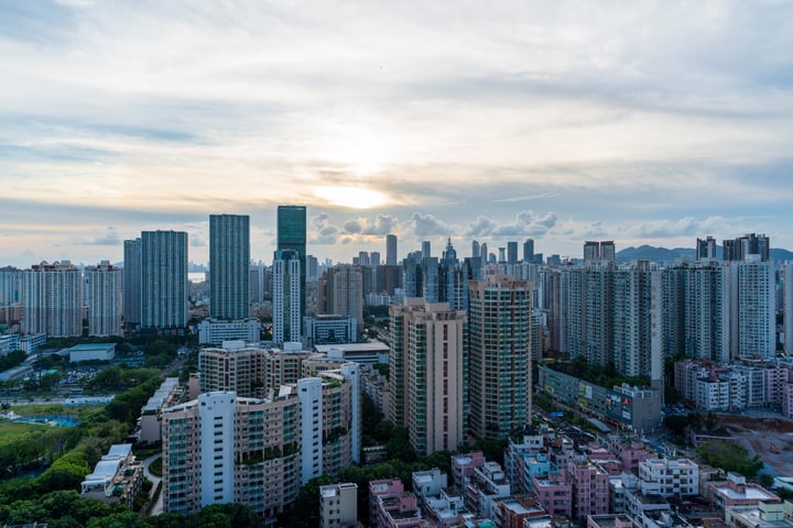A cityscape with tall buildings, including skyscrapers, is captured during sunset, with the sun setting behind the buildings and casting a warm glow. The sky is filled with clouds, and the sun is partially obscured by the clouds. The city is densely populated with buildings, and a pool is visible in the foreground.