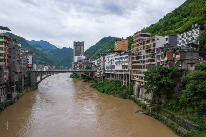 A serene mountain landscape with a river flowing through it, surrounded by buildings with varying architectural styles, and a bridge connecting the two sides.