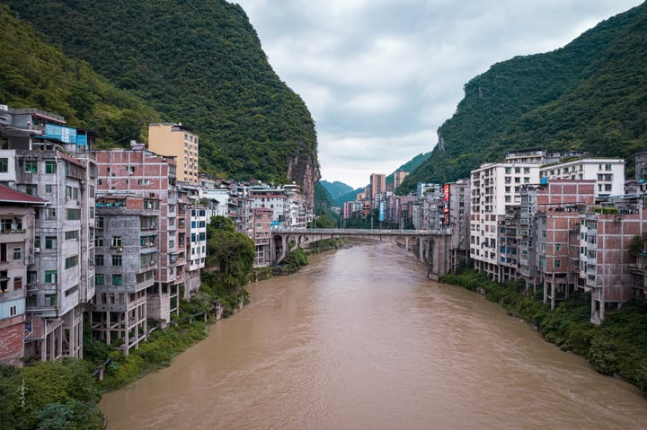 A serene cityscape with a river, a bridge, and colorful buildings, surrounded by lush green mountains.