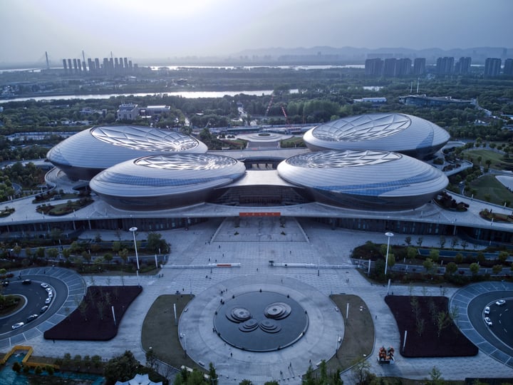 An aerial view of a modern sports complex with two circular domes, surrounded by a large circular courtyard and a fountain, with a city skyline in the background.