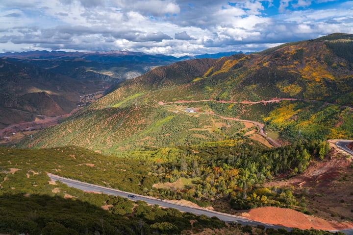 A panoramic view of a mountainous landscape with a winding road, surrounded by lush greenery and a blue sky with white clouds.