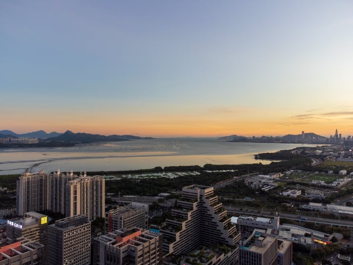 A cityscape at sunset, with a large body of water reflecting the sky, tall buildings, and mountains in the background.