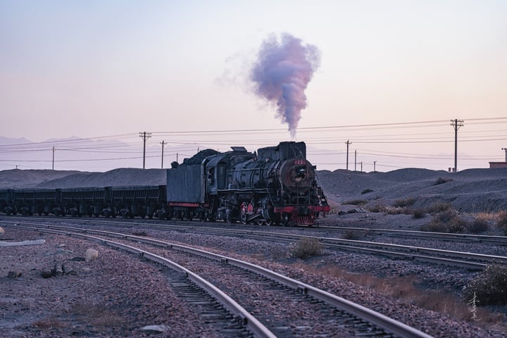 A black steam locomotive pulls a line of black cars on a curved track, with power lines overhead and a mountain in the distance.