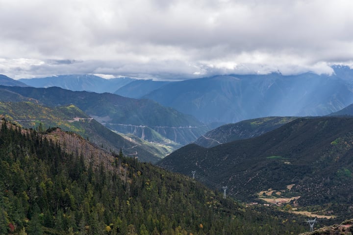 A mountainous valley with a winding road, lush green trees, and a cloudy sky.