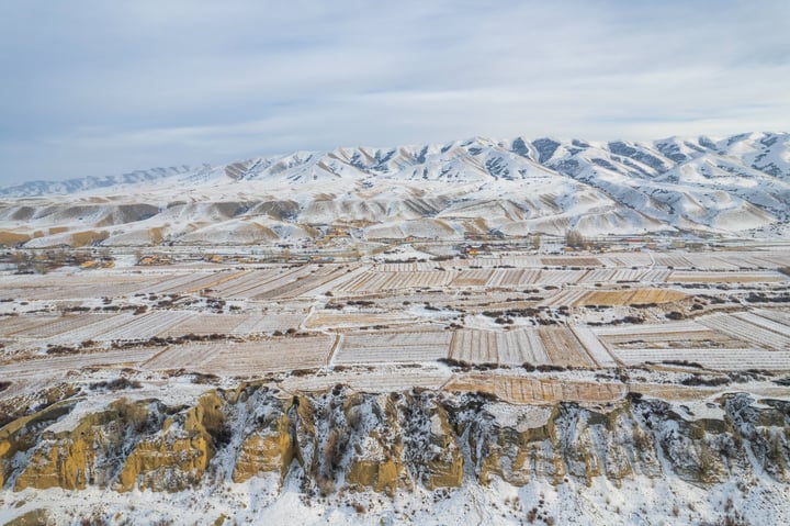 A snowy mountain landscape with a valley, fields, and houses, with a cloudy sky above.