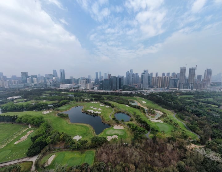 An aerial view of a golf course surrounded by a large body of water, with a city skyline in the background.