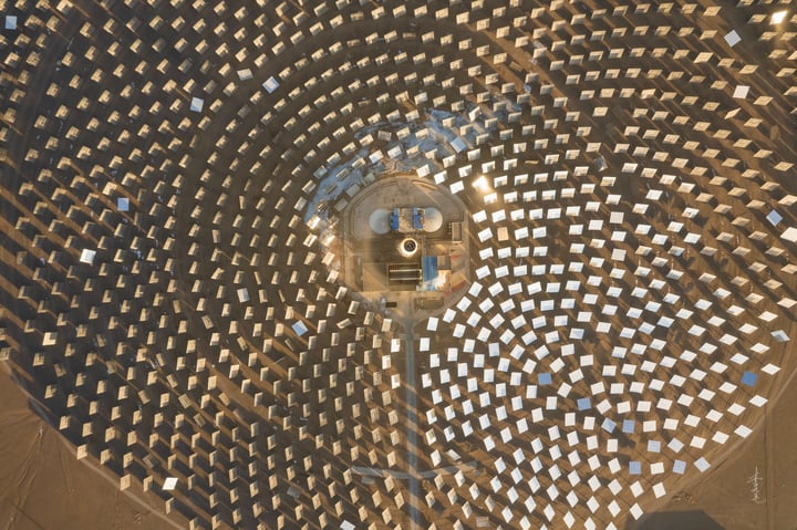 An aerial view of a large solar farm in a desert-like environment, with solar panels arranged in a circular pattern and a large white sun tower in the center. The farm is surrounded by a vast expanse of sandy terrain and a few wind turbines in the distance.