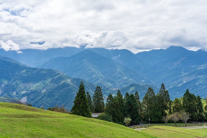 A serene mountain landscape with lush green hills, trees, and a clear blue sky with white clouds.