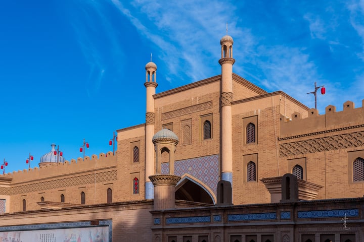 A large beige building with multiple minarets and towers, adorned with flags, stands against a clear blue sky.