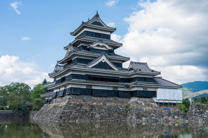 The image features the majestic Himeiji Castle, a Japanese landmark, situated on a stone wall by a body of water. The castle's black and white facade contrasts with the surrounding greenery, and the clear blue sky with white clouds adds to the serene atmosphere. The castle is surrounded by lush trees and mountains, creating a picturesque scene.