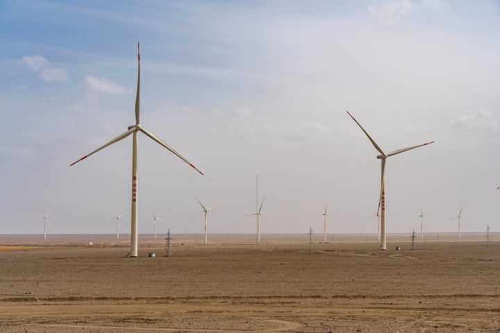 A vast desert landscape with a row of white wind turbines, each with red and white striped poles, under a clear blue sky.