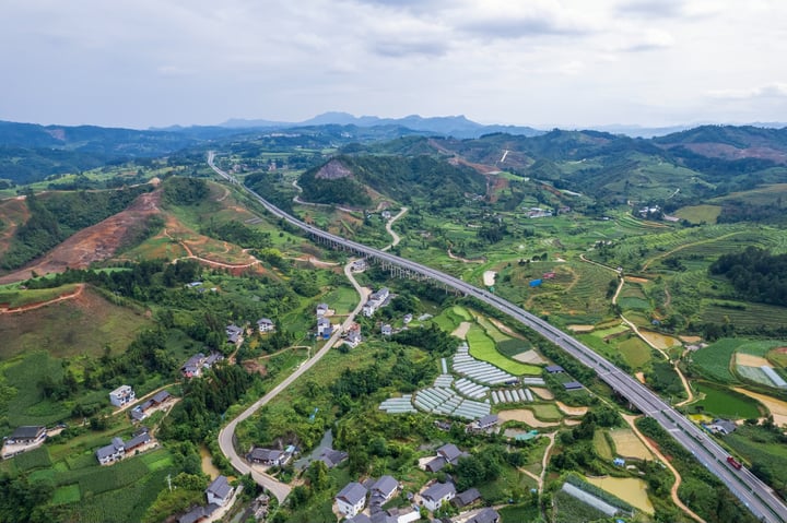 An aerial view of a rural area with a winding highway, surrounded by lush green fields and houses, with mountains in the distance.