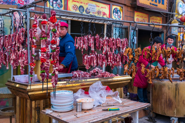 A bustling market scene features a vendor selling meat, a man in a red hat, and a woman in a red jacket. The market has a wooden table with white plates and bowls, and a yellow sign with Chinese characters. The background shows a building with a red awning and a yellow sign.