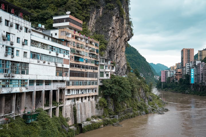 A cityscape with tall buildings on a cliff overlooking a river, surrounded by lush greenery and a cloudy sky.