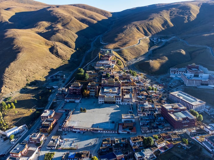 An aerial view of a mountainous region with a large, multi-level building in the center, surrounded by a mix of brown and green hills. The buildings are predominantly white and red, with some green roofs. The sky is clear blue, and the sun shines brightly.