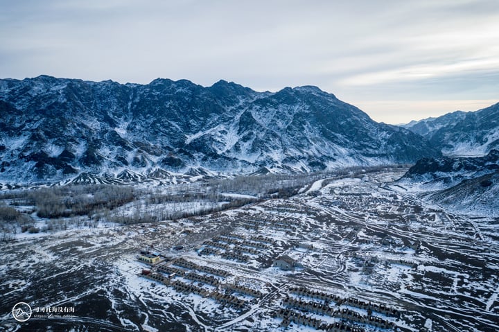 A snowy mountain range with a valley, bare trees, and a clear blue sky.