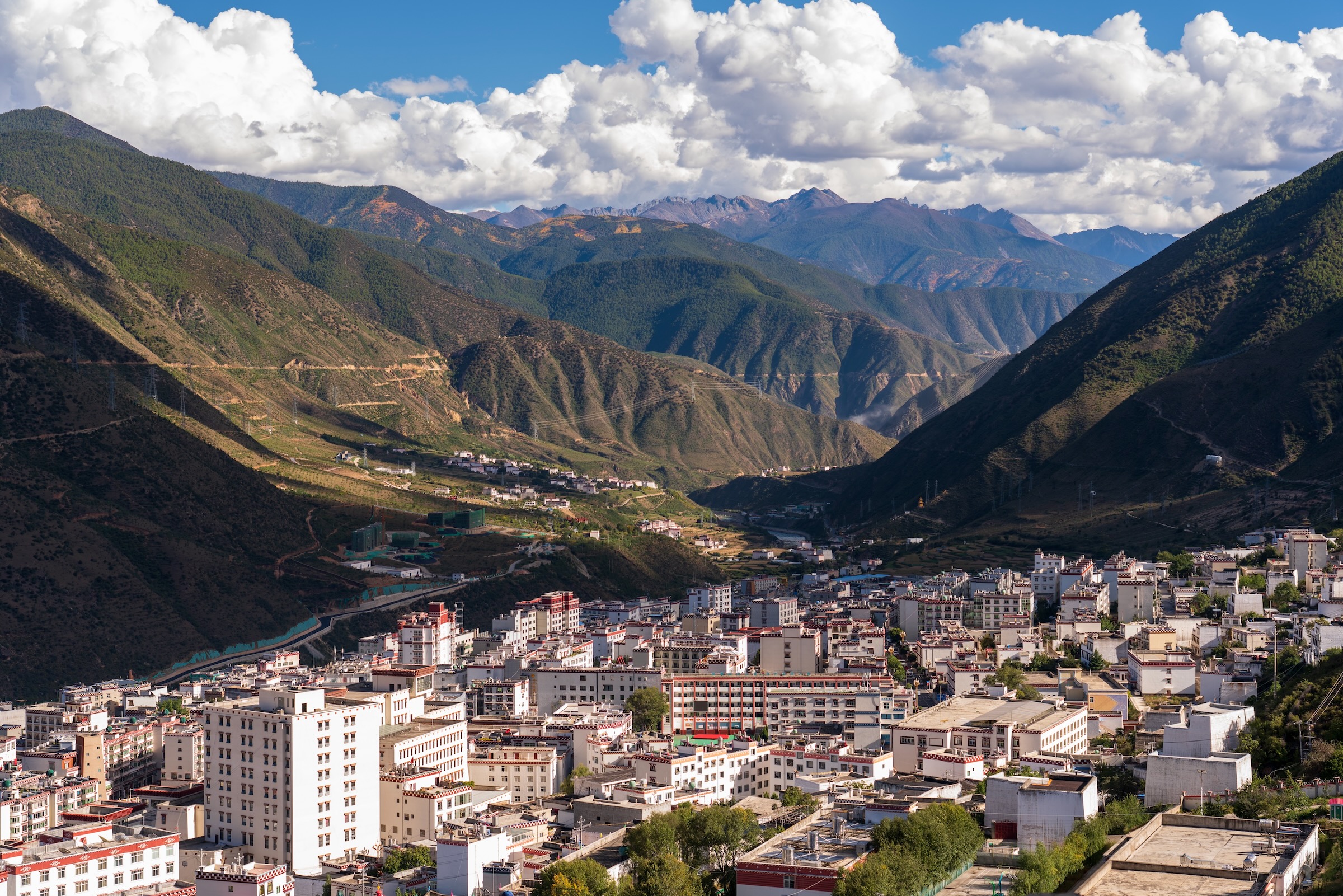 A panoramic view of a mountainous city nestled between verdant mountains, with a winding river and a clear blue sky.
