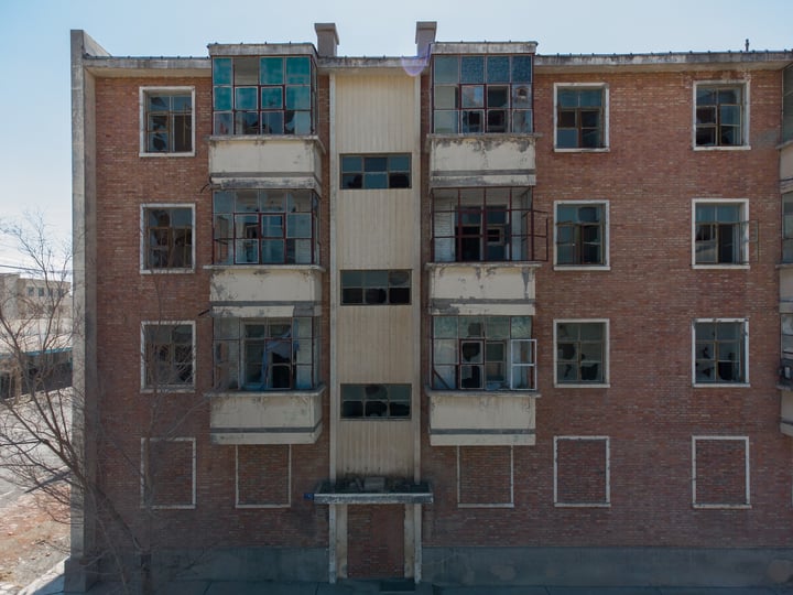 A large, multi-story brick building with numerous windows and balconies is in a state of disrepair, with some windows missing and others broken. The building is surrounded by trees and a parking lot.