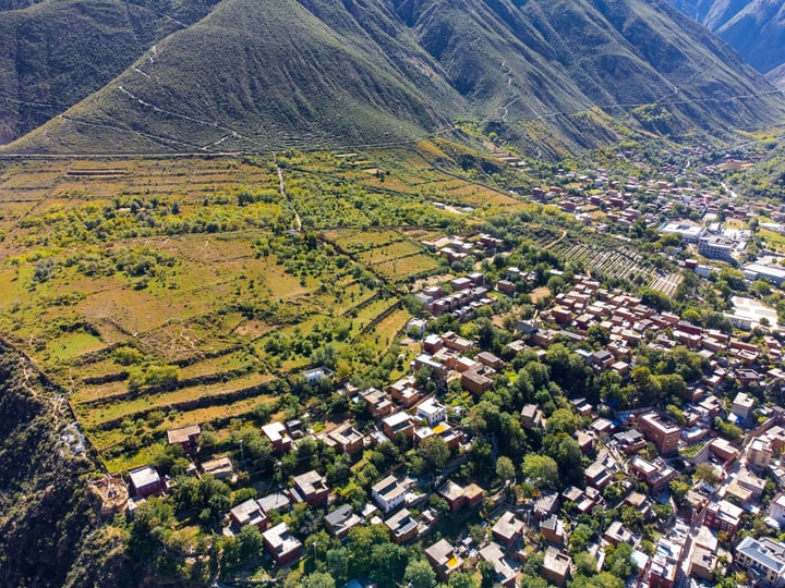 An aerial view of a small village nestled in a valley, surrounded by lush green mountains, with red and brown buildings scattered across the landscape.
