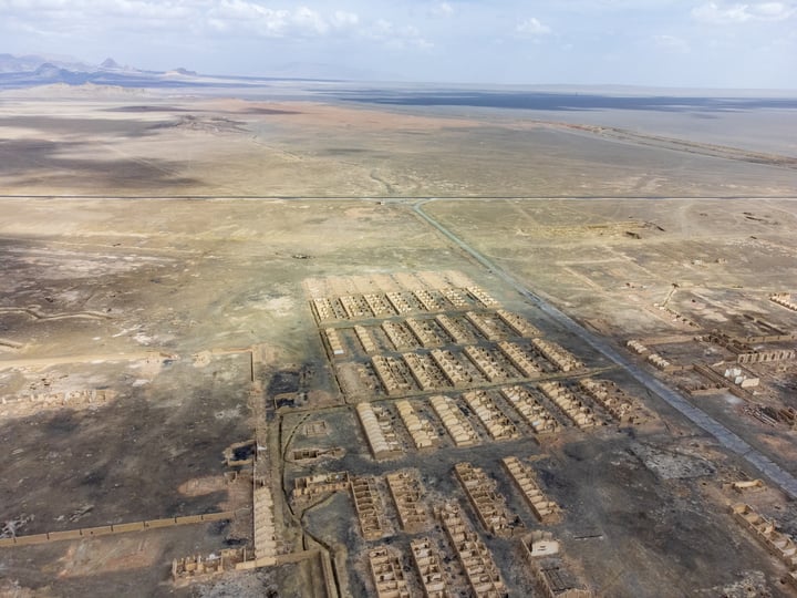 An aerial view of a desert landscape with a grid-like layout of buildings and structures, surrounded by sandy terrain and distant mountains.