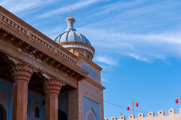A large, ornate building with a blue dome and golden spire stands against a clear blue sky, adorned with red flags. The building features intricate brickwork and columns, and is located on a street with power lines.