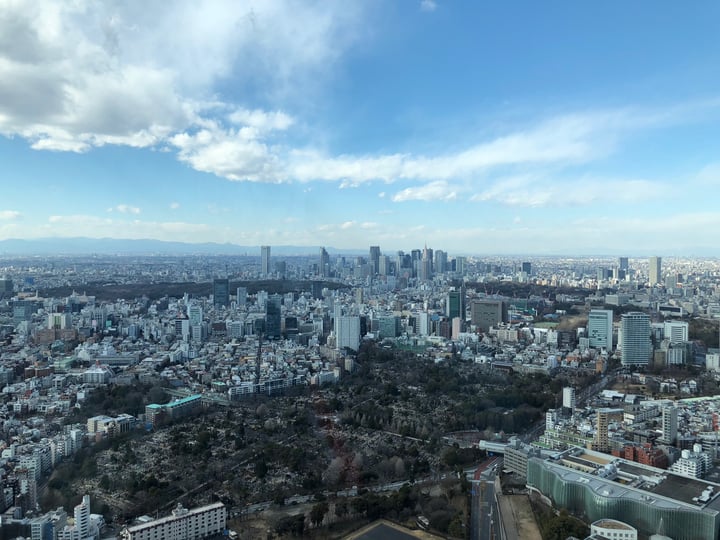 A bustling cityscape with towering skyscrapers and a bridge spanning over a river is captured from a high vantage point, showcasing the urban layout and the vibrant greenery of parks and parks.