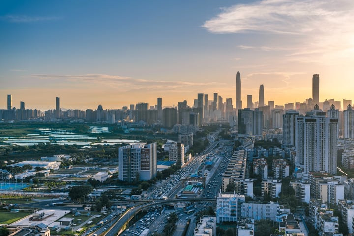A cityscape at sunset, with tall buildings in the background, a busy highway, and a large body of water in the foreground.