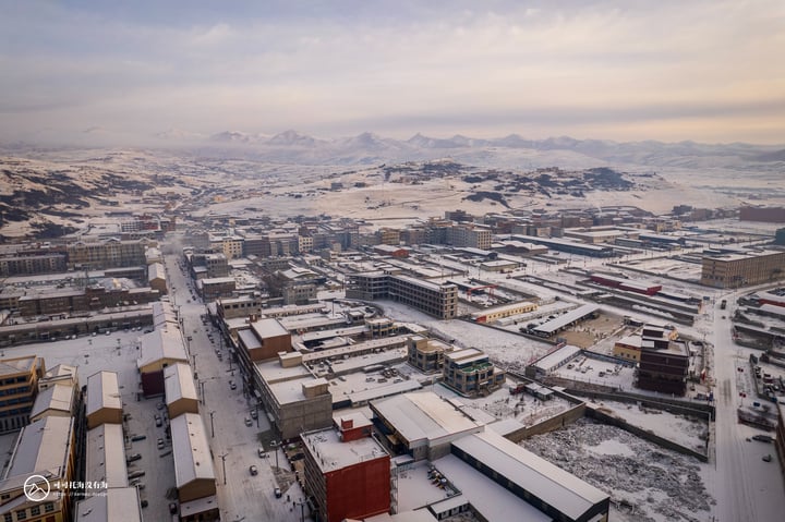 An aerial view of a snowy city with a mix of buildings, roads, and mountains in the background.