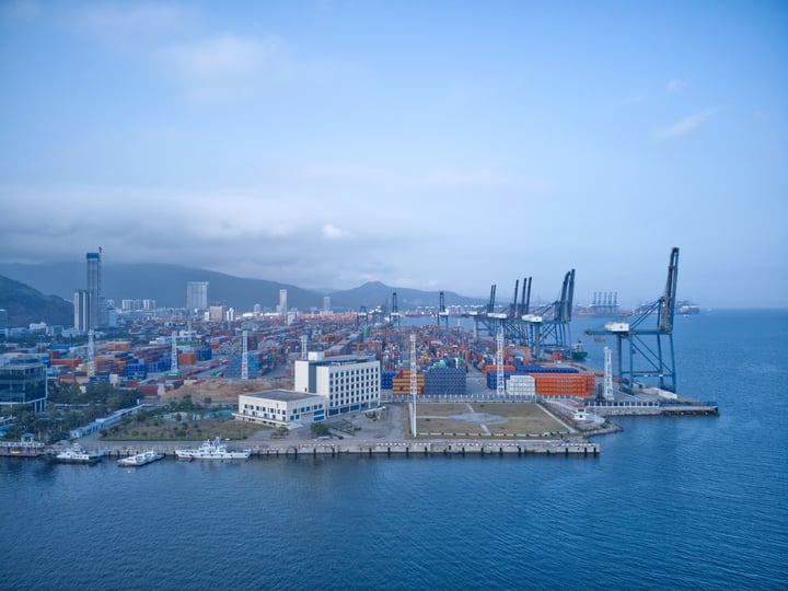 An aerial view of a bustling port with a large number of containers, cranes, and boats, surrounded by mountains and a clear blue sky.