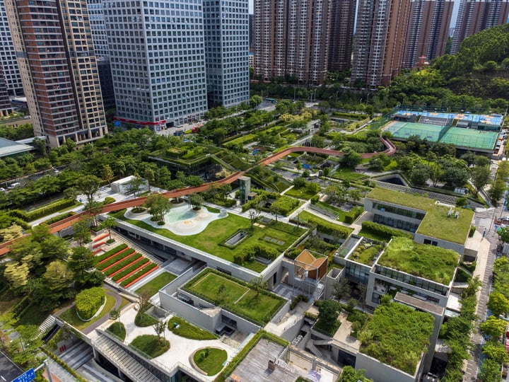 An aerial view of a well-planned urban park with a variety of green spaces, a water feature, and a basketball court, surrounded by tall buildings and a clear blue sky.