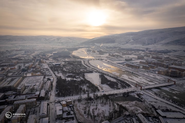 An aerial view of a snowy mountain town with a winding river, a bridge, and a few buildings, all covered in snow.
