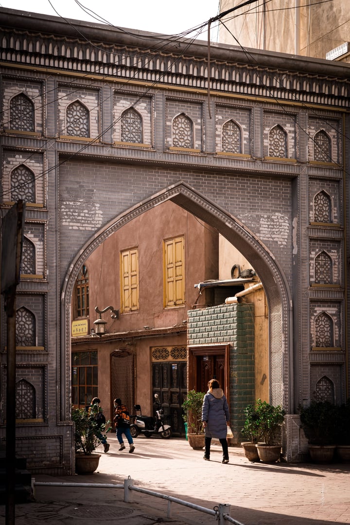 A man in a blue coat walks towards a large archway in an old town, surrounded by buildings with intricate designs and potted plants.