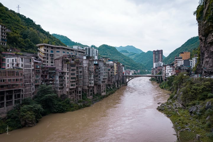 A cityscape on a hillside, with tall buildings of varying heights and colors, surrounded by lush greenery. The river, a brown, choppy body, flows through the city, reflecting the overcast sky.