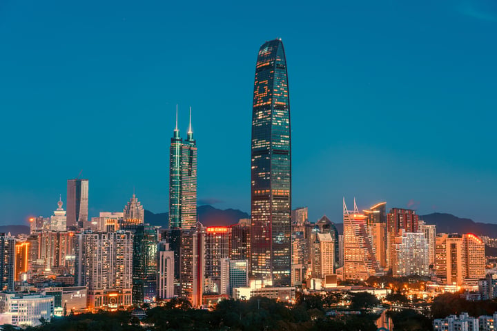 The image captures a breathtaking view of Hong Kong's skyline at dusk, with the iconic World Trade Center tower prominently displayed. The cityscape is illuminated by warm lights, creating a vibrant contrast against the deep blue sky. The mountains in the distance add a touch of nature to the urban landscape. The image is taken from a low angle, emphasizing the towering skyscrapers and the city's grandeur.