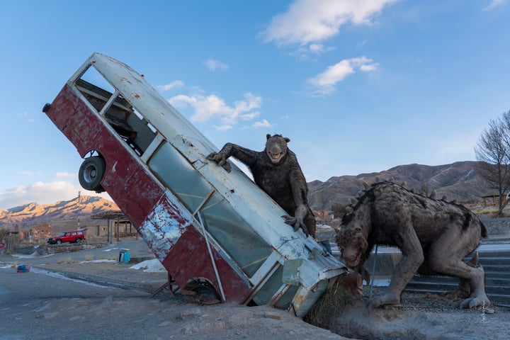 A man is leaning over a broken car, while a wolf stands nearby, both on a road with mountains in the background.