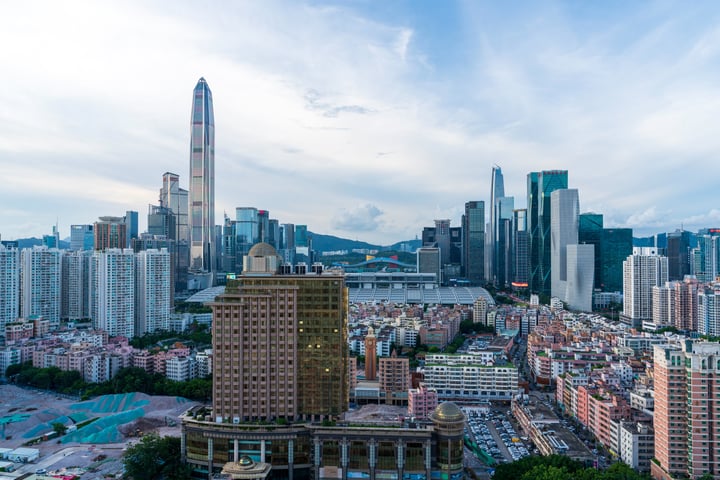 A panoramic view of a bustling city with tall buildings, a skyscraper, and a bridge, surrounded by mountains and a clear blue sky.