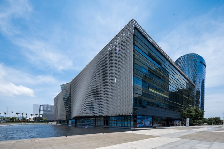 The image features a modern, curved building with a large glass facade, situated by a body of water. The building's design is modern and sleek, with a curved shape and a large glass window. The building is situated on a sidewalk, with a few trees nearby. The sky is clear and blue, with a few clouds scattered across it. The building's location by the water provides a serene and picturesque setting.